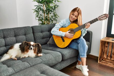 Téléchargez les photos : Jeune femme caucasienne jouant de la guitare classique assise sur un canapé avec chien à la maison - en image libre de droit