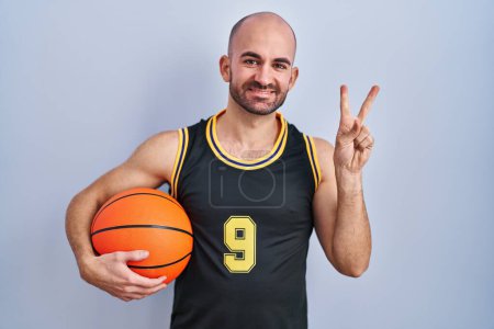 Téléchargez les photos : Jeune homme chauve avec barbe portant un uniforme de basket-ball tenant la balle souriant en regardant la caméra montrant les doigts faire signe de victoire. numéro deux. - en image libre de droit