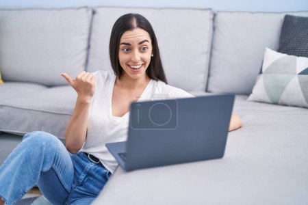 Téléchargez les photos : Jeune femme brune utilisant un ordinateur portable à la maison pointant le pouce vers le côté souriant heureux avec la bouche ouverte - en image libre de droit