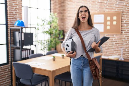 Photo for Young brunette woman working at the office holding bike helmet angry and mad screaming frustrated and furious, shouting with anger looking up. - Royalty Free Image