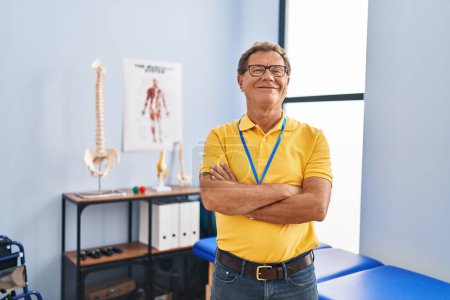 Foto de Hombre de mediana edad fisioterapeuta sonriendo confiado de pie con los brazos cruzados gesto en la clínica de fisioterapia - Imagen libre de derechos