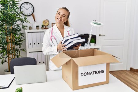 Téléchargez les photos : Young blonde girl wearing doctor uniform putting book in donations box at clinic - en image libre de droit