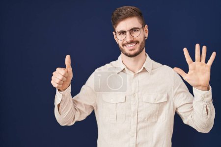 Foto de Hombre hispano con barba de pie sobre fondo azul mostrando y señalando hacia arriba con los dedos número seis mientras sonríe confiado y feliz. - Imagen libre de derechos