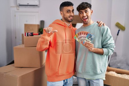 Téléchargez les photos : Young hispanic gay couple moving to a new home holding banknotes pointing thumb up to the side smiling happy with open mouth - en image libre de droit