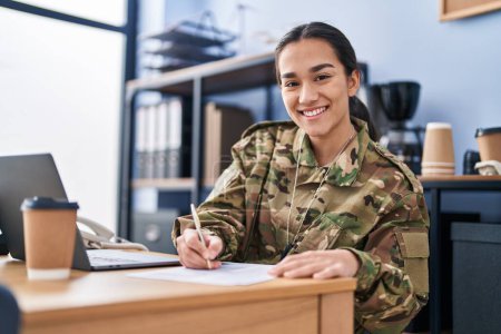Téléchargez les photos : Jeune femme hispanique soldat de l'armée en utilisant l'écriture d'ordinateur portable sur le document au bureau - en image libre de droit