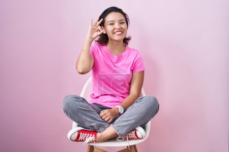 Photo for Hispanic young woman sitting on chair over pink background smiling positive doing ok sign with hand and fingers. successful expression. - Royalty Free Image