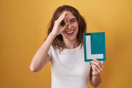 Photo for Beautiful brunette woman holding l sign for new driver smiling happy doing ok sign with hand on eye looking through fingers - Royalty Free Image
