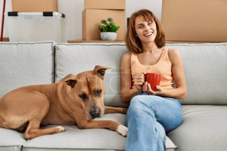 Photo for Young caucasian woman drinking coffee sitting on sofa with dog at home - Royalty Free Image