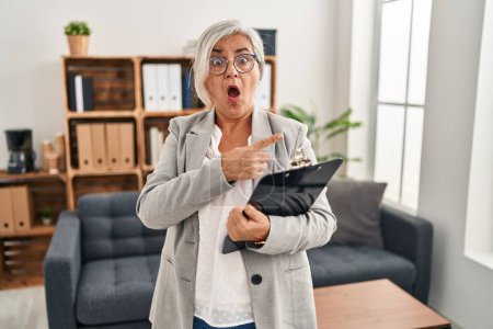 Foto de Middle age woman with grey hair at consultation office surprised pointing with finger to the side, open mouth amazed expression. - Imagen libre de derechos