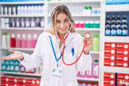 Foto de Young blonde woman working at pharmacy drugstore holding stethoscope celebrating achievement with happy smile and winner expression with raised hand - Imagen libre de derechos
