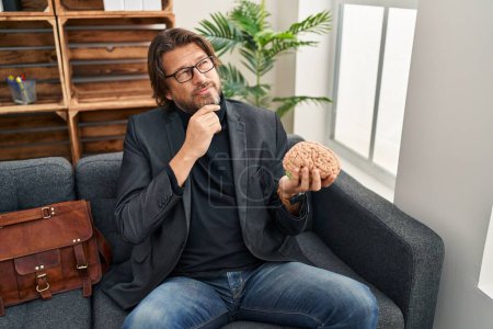 Photo for Handsome middle age man at consultation office holding brain serious face thinking about question with hand on chin, thoughtful about confusing idea - Royalty Free Image