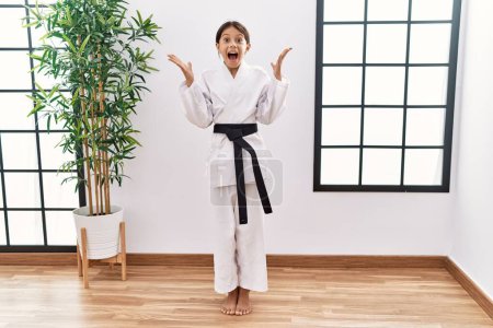 Téléchargez les photos : Young hispanic girl wearing karate kimono and black belt celebrating crazy and amazed for success with arms raised and open eyes screaming excited. winner concept - en image libre de droit