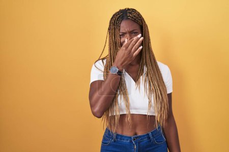 Téléchargez les photos : African american woman with braided hair standing over yellow background smelling something stinky and disgusting, intolerable smell, holding breath with fingers on nose. bad smell - en image libre de droit