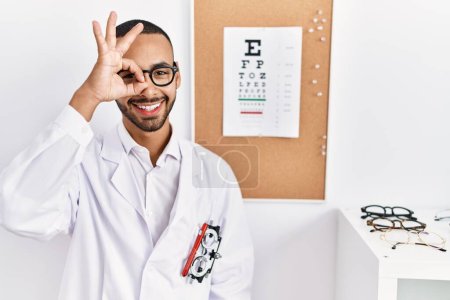 Téléchargez les photos : African american optician man standing by eyesight test doing ok gesture with hand smiling, eye looking through fingers with happy face. - en image libre de droit