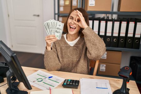 Foto de Young beautiful woman working at small business ecommerce holding money smiling happy doing ok sign with hand on eye looking through fingers - Imagen libre de derechos