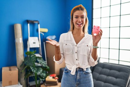 Foto de Young blonde woman moving to a new home holding piggy bank celebrating achievement with happy smile and winner expression with raised hand - Imagen libre de derechos