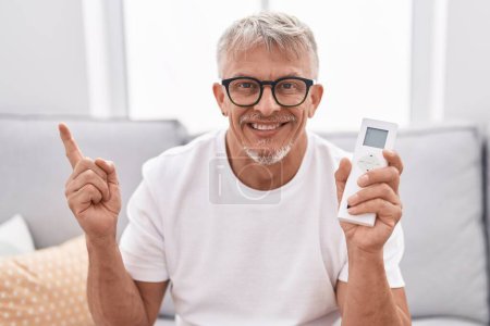 Foto de Hispanic man with grey hair holding air conditioner control smiling happy pointing with hand and finger to the side - Imagen libre de derechos
