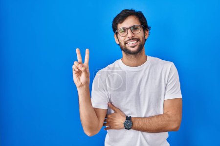 Photo for Handsome latin man standing over blue background smiling with happy face winking at the camera doing victory sign. number two. - Royalty Free Image