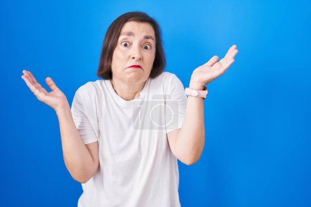 Photo for Middle age hispanic woman standing over blue background clueless and confused expression with arms and hands raised. doubt concept. - Royalty Free Image