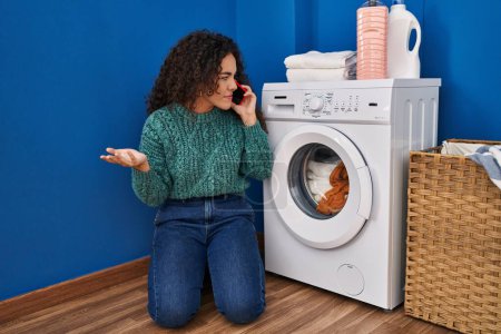 Téléchargez les photos : Jeune belle femme hispanique parlant sur smartphone avec technicien pour machine à laver cassée à la buanderie - en image libre de droit