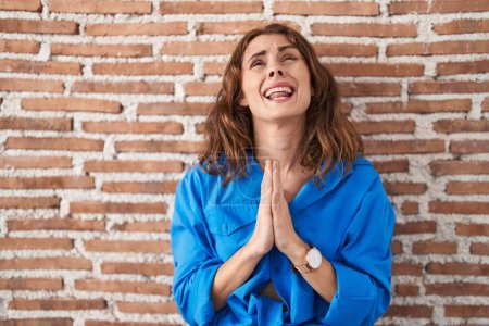 Photo for Beautiful brunette woman standing over bricks wall begging and praying with hands together with hope expression on face very emotional and worried. begging. - Royalty Free Image