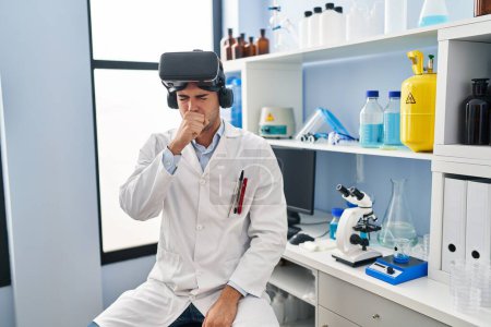 Foto de Young hispanic man working at scientist laboratory wearing vr glasses feeling unwell and coughing as symptom for cold or bronchitis. health care concept. - Imagen libre de derechos