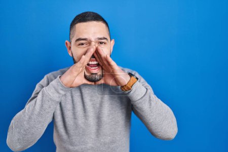 Téléchargez les photos : Homme hispanique debout sur fond bleu criant à haute voix avec les mains sur la bouche - en image libre de droit