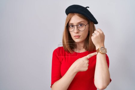 Foto de Young redhead woman standing wearing glasses and beret in hurry pointing to watch time, impatience, looking at the camera with relaxed expression - Imagen libre de derechos