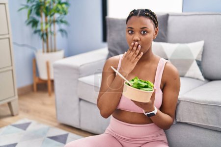 Foto de Mujer afroamericana con trenzas comiendo ensalada después de hacer ejercicio en casa cubriendo la boca con la mano, conmocionada y temerosa de equivocarse. expresión sorprendida - Imagen libre de derechos