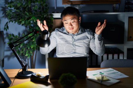 Photo for Young chinese man working using computer laptop at night clueless and confused expression with arms and hands raised. doubt concept. - Royalty Free Image