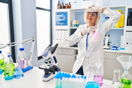 Téléchargez les photos : Middle age blonde woman working at scientist laboratory touching forehead for illness and fever, flu and cold, virus sick - en image libre de droit