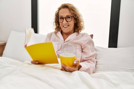 Photo for Middle age caucasian woman reading book and drinking coffee sitting on the bed at bedroom. - Royalty Free Image