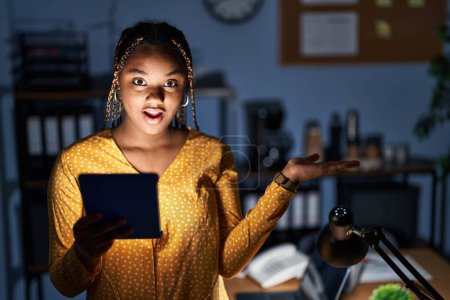 Téléchargez les photos : Femme afro-américaine avec des tresses travaillant au bureau la nuit avec tablette pointant vers le côté avec les mains ouvertes paumes montrant l'espace de copie, présentant la publicité souriant heureux excité - en image libre de droit