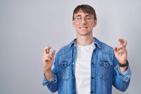 Photo for Caucasian blond man standing wearing glasses gesturing finger crossed smiling with hope and eyes closed. luck and superstitious concept. - Royalty Free Image
