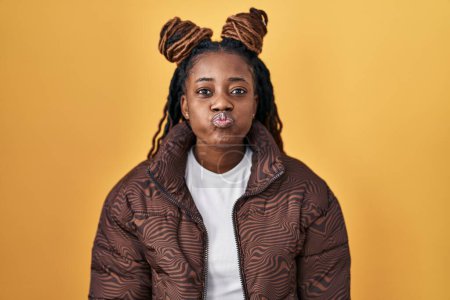 Téléchargez les photos : Femme africaine avec des cheveux tressés debout sur fond jaune joues gonflées avec un visage drôle. bouche gonflée d'air, expression folle. - en image libre de droit
