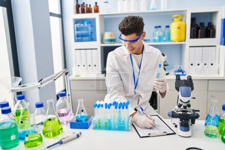 Photo for Young hispanic man wearing scientist uniform writing on clipboard at laboratory - Royalty Free Image