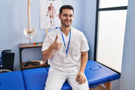 Téléchargez les photos : Young hispanic man with beard working at pain recovery clinic doing happy thumbs up gesture with hand. approving expression looking at the camera showing success. - en image libre de droit