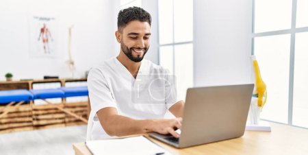 Photo for Young arab man wearing physiotherapist uniform using laptop at clinic - Royalty Free Image