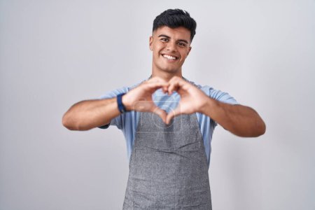 Foto de Joven hispano vistiendo delantal sobre fondo blanco sonriendo en amor haciendo forma de símbolo del corazón con las manos. concepto romántico. - Imagen libre de derechos
