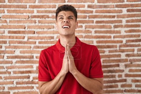 Photo for Young hispanic man standing over bricks wall begging and praying with hands together with hope expression on face very emotional and worried. begging. - Royalty Free Image