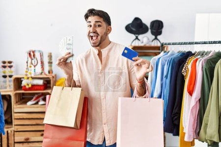 Foto de Joven hombre hispano sosteniendo bolsas de compras y tarjeta de crédito celebrando loco y sorprendido por el éxito con los ojos abiertos gritando emocionado. - Imagen libre de derechos