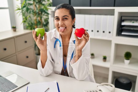 Téléchargez les photos : Young hispanic woman working at dietitian clinic holding green apple sticking tongue out happy with funny expression. - en image libre de droit