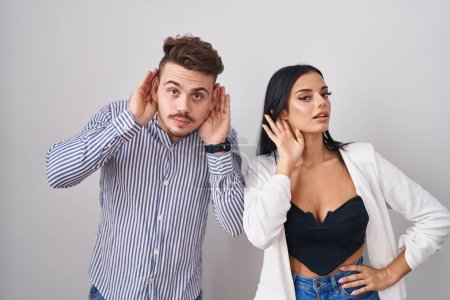Photo for Young hispanic couple standing over white background trying to hear both hands on ear gesture, curious for gossip. hearing problem, deaf - Royalty Free Image