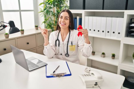 Photo for Hispanic doctor woman holding support red ribbon smiling happy pointing with hand and finger to the side - Royalty Free Image
