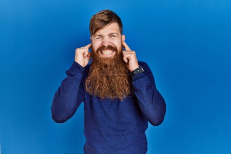 Foto de Redhead man with long beard wearing casual blue sweater over blue background covering ears with fingers with annoyed expression for the noise of loud music. deaf concept. - Imagen libre de derechos
