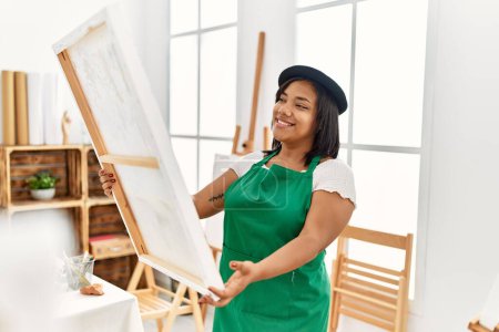 Téléchargez les photos : Hispanic brunette woman holding canvas proud of painting at art studio - en image libre de droit