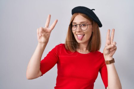Téléchargez les photos : Jeune femme rousse debout portant des lunettes et un béret souriant avec la langue montrant les doigts des deux mains faisant signe de victoire. numéro deux. - en image libre de droit
