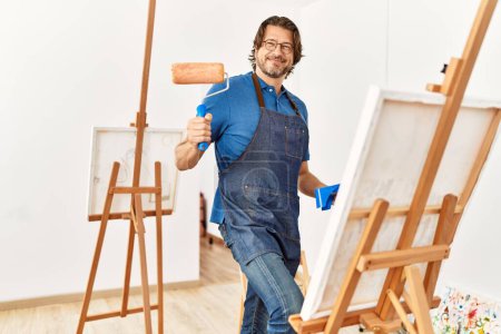Foto de Edad media hombre caucásico sonriendo dibujo seguro utilizando rodillo de pintura en el estudio de arte - Imagen libre de derechos