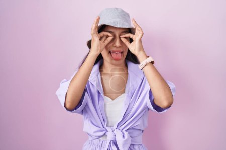 Téléchargez les photos : Young hispanic woman standing over pink background wearing hat doing ok gesture like binoculars sticking tongue out, eyes looking through fingers. crazy expression. - en image libre de droit