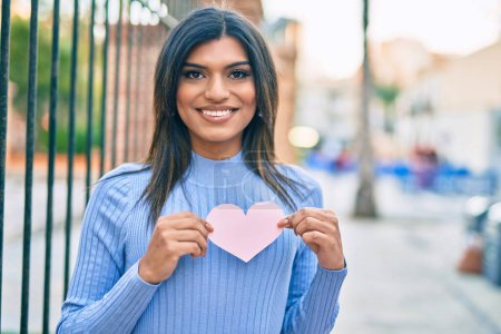 Photo for Beautiful hispanic woman smiling holding pink heart at the city - Royalty Free Image
